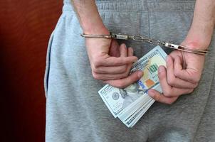 Ein festgenommener Typ in grauer Hose mit gefesselten Händen hält eine riesige Menge Dollarscheine. Rückansicht foto