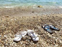 Auf den Steinen stehen gummiweiße Damen- und blaue Herrenpantoffeln. Urlaub auf See, Reise in ein heißes und warmes Land. Schuhe sind am Strand, während eine Person schwimmt foto