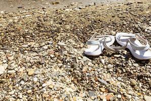 Auf den Steinen stehen weiße Gummipantoffeln. Urlaub auf See, Reise in ein heißes und warmes Land. Schuhe sind am Strand, während eine Person schwimmt foto