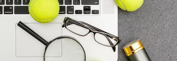 draufsicht auf laptop, sportgeräte, tennisball, federball, brille auf dem grauen tisch der sportverwaltung. geschäftskonzept. foto