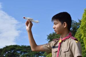 asiatischer pfadfinder hält weißes aroplane-modell vor bewölktem und blauem hintergrund, weicher und selektiver fokus. foto