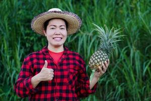 asiatische bäuerin trägt hut, rotes kariertes hemd, hält ananasfrucht, daumen hoch. konzept, landwirtschaftsernte in thailand. Bauer zufrieden. Bio-Pflanzen. saisonale Früchte. foto