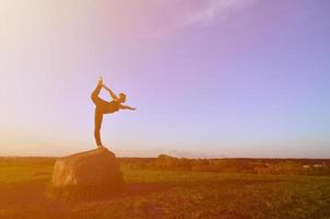 Silhouette eines jungen blonden Mädchens im Sportanzug Yoga auf einem malerischen grünen Hügel am Abend bei Sonnenuntergang. das konzept der übung und des gesunden lebensstils foto