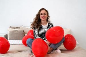 Valentinstag. junge glückliche frau, die auf dem bett mit roten herzförmigen luftballons sitzt foto