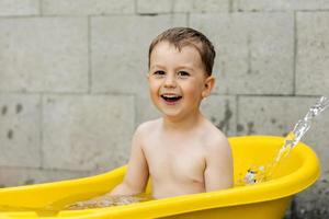 Süßer kleiner Junge, der im Freien in der gelben Wanne badet. glückliches kind plantscht, spielt mit wasser und hat spaß. Sommersaison und Erholung. Kühl bleiben in der Sommerhitze. Wasserspaß im Hinterhof. foto