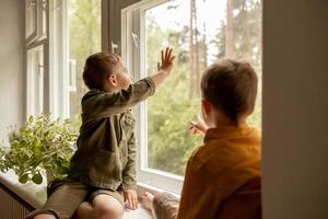 Kinder sitzen auf der Fensterbank und warten darauf, dass jemand kommt. zwei Brüder, Freunde. süße vorschulkinder allein zu hause. Jungen warten auf ihre Mutter oder ihren Vater. Einsamkeit. beschäftigte Eltern. foto