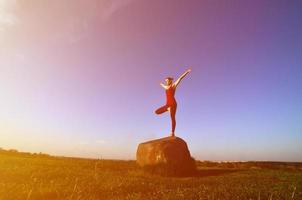 Silhouette eines jungen blonden Mädchens im Sportanzug Yoga auf einem malerischen grünen Hügel am Abend bei Sonnenuntergang. das konzept der übung und des gesunden lebensstils foto