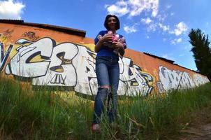 Porträt eines emotionalen jungen Mädchens mit schwarzen Haaren und Piercings. Ein Weitwinkelfoto eines Mädchens mit Aerosol-Farbdosen in den Händen auf einem Graffiti-Wandhintergrund. ein modernes Porträt eines Fisheye-Objektivs foto