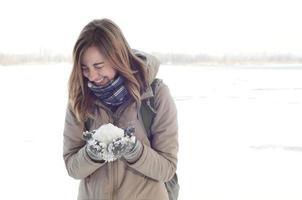 Ein junges und fröhliches kaukasisches Mädchen in einem braunen Mantel hält im Winter einen Schneeball im Hintergrund einer Horizontlinie zwischen dem Himmel und einem zugefrorenen See foto