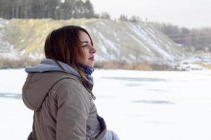 Ein junges kaukasisches Mädchen in einem braunen Mantel, das im Winter in die Ferne auf der Horizontlinie zwischen dem Himmel und dem zugefrorenen See starrt foto