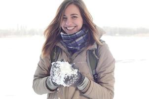 Ein junges und fröhliches kaukasisches Mädchen in einem braunen Mantel hält im Winter einen Schneeball im Hintergrund einer Horizontlinie zwischen dem Himmel und einem zugefrorenen See foto