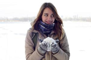 Ein junges und fröhliches kaukasisches Mädchen in einem braunen Mantel hält im Winter einen Schneeball im Hintergrund einer Horizontlinie zwischen dem Himmel und einem zugefrorenen See foto