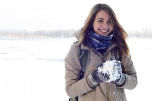 Ein junges und fröhliches kaukasisches Mädchen in einem braunen Mantel hält im Winter einen Schneeball im Hintergrund einer Horizontlinie zwischen dem Himmel und einem zugefrorenen See foto