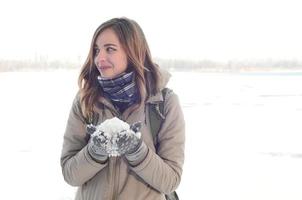 Ein junges und fröhliches kaukasisches Mädchen in einem braunen Mantel hält im Winter einen Schneeball im Hintergrund einer Horizontlinie zwischen dem Himmel und einem zugefrorenen See foto