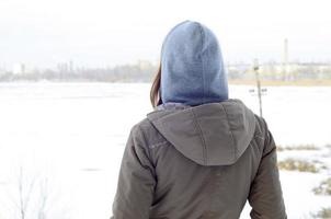 Ein junges kaukasisches Mädchen in einem braunen Mantel, das im Winter in die Ferne auf der Horizontlinie zwischen dem Himmel und dem zugefrorenen See starrt foto