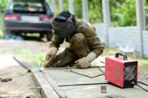 alter schweißer in brauner uniform, schweißmaske und schweißleder, schweißmetalltür mit lichtbogenschweißgerät im freien foto