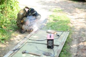 alter schweißer in brauner uniform, schweißmaske und schweißleder, schweißmetalltür mit lichtbogenschweißgerät im freien foto