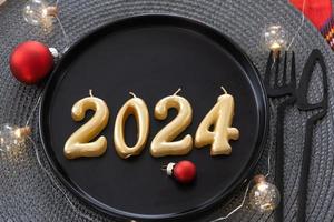 goldene figuren 2024 aus kerzen auf einem schwarzen teller auf einem festlich gedeckten tisch mit einer neujahrsportion. Einrichtung im Loft-Stil, Party, Fest. Korbserviette, Gabel, Messer foto