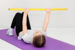 Kleines Mädchen liegt auf einer Matte und hält einen Gymnastikstock auf ausgestreckten Armen, Kinderfitness, Sport foto