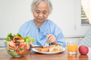 asiatische Seniorin oder ältere Patientin der alten Dame, die Lachssteakfrühstück mit gesundem Gemüse isst, während sie im Krankenhaus sitzt und hungrig auf dem Bett liegt. foto