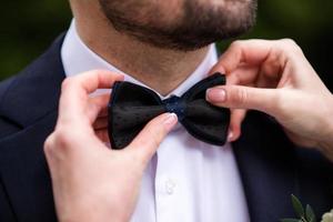 die Hände der Fliege des Bräutigams. Stilvolle Trauzeugen helfen dem glücklichen Bräutigam, sich morgens für die Hochzeitszeremonie fertig zu machen. Luxusmann im Anzug im Zimmer. Hochzeitstag. foto
