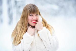schöne lächelnde selbstbewusste junge weiße frau hübsches gesicht mit leuchtend roten lippen im weißen pelzmantel, der in die kamera blickt, die allein im verschneiten wald posiert foto