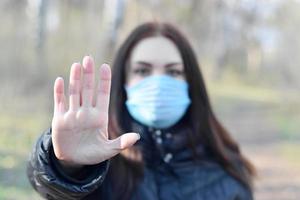 Porträt einer jungen brünetten Frau in blauer Schutzmaske zeigt Stoppgeste im Freien im Frühlingsholz foto