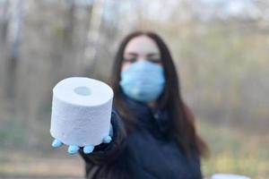 Covidiotisches Konzept. Junge Frau in Schutzmaske gibt Rolle Toilettenpapier im Freien im Frühlingsholz foto