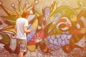 Foto eines jungen Mannes in Jeansshorts und weißem Hemd. Der Typ zeichnet auf die Graffitiwand eine Zeichnung mit Aerosolfarben in verschiedenen Farben. das Konzept des Rowdytums und der Sachbeschädigung