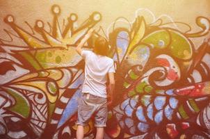 Foto eines jungen Mannes in Jeansshorts und weißem Hemd. Der Typ zeichnet auf die Graffitiwand eine Zeichnung mit Aerosolfarben in verschiedenen Farben. das Konzept des Rowdytums und der Sachbeschädigung