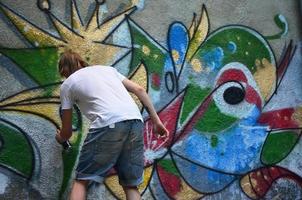 Foto eines jungen Mannes in Jeansshorts und weißem Hemd. Der Typ zeichnet auf die Graffitiwand eine Zeichnung mit Aerosolfarben in verschiedenen Farben. das Konzept des Rowdytums und der Sachbeschädigung