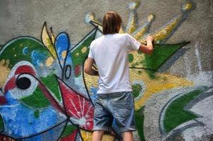 Foto eines jungen Mannes in Jeansshorts und weißem Hemd. Der Typ zeichnet auf die Graffitiwand eine Zeichnung mit Aerosolfarben in verschiedenen Farben. das Konzept des Rowdytums und der Sachbeschädigung