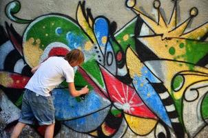 Foto eines jungen Mannes in Jeansshorts und weißem Hemd. Der Typ zeichnet auf die Graffitiwand eine Zeichnung mit Aerosolfarben in verschiedenen Farben. das Konzept des Rowdytums und der Sachbeschädigung