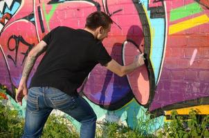 Der junge Graffiti-Künstler mit Rucksack und Gasmaske am Hals malt bunte Graffiti in rosa Tönen auf die Ziegelwand. Street Art und zeitgenössischer Malprozess foto