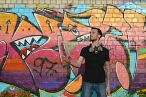 Der junge Graffiti-Künstler mit Gasmaske am Hals wirft seine Sprühdose gegen bunte rosafarbene Graffiti an der Ziegelwand. Street Art und zeitgenössischer Malprozess foto