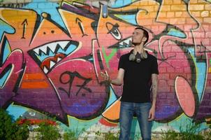 Der junge Graffiti-Künstler mit Gasmaske am Hals wirft seine Sprühdose gegen bunte rosafarbene Graffiti an der Ziegelwand. Street Art und zeitgenössischer Malprozess foto