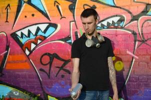 junger kaukasischer graffiti-künstler in schwarzem t-shirt mit silberner spraydose in der nähe von bunten graffiti in rosa tönen an der mauer. Street Art und zeitgenössischer Malprozess foto