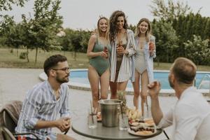 Gruppe junger Leute, die mit Getränken jubeln und Obst am Pool im Garten essen foto