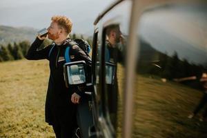 junger Mann entspannt und trinkt Kaffee an der Motorhaube des Geländefahrzeugs auf dem Land foto