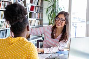 junge berufstätige arbeiten im unternehmen mit laptop und tablet. zwei frauen sitzen am tisch, unterhalten sich lächelnd, schauen auf den bildschirm eines silbernen pc und halten analysen in den händen. foto