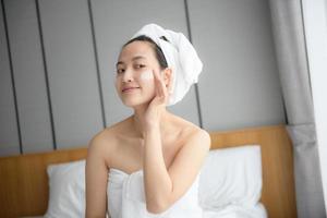 glückliche junge asiatin, die gesichtslotionen aufträgt, während sie ein handtuch trägt und ihr gesicht berührt. tägliches Make-up und Hautpflege foto