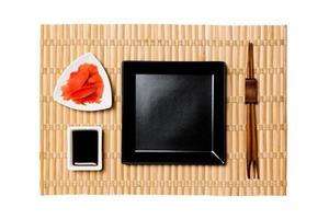 leere schwarze quadratische Platte mit Essstäbchen für Sushi, Ingwer und Sojasauce auf gelbem Bambusmattenhintergrund. Draufsicht mit Kopierbereich für Ihr Design foto