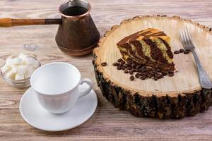 Ein Schokoladenkuchen auf Holzstumpf mit Kaffeetasse, Gabel, Kaffeebohnen und Schüssel mit Zuckerwürfeln auf hellem Holzhintergrund foto