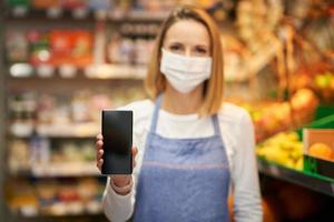 Verkäuferin in medizinischer Maske, die Smartphone gegen Lebensmittelgeschäft hält