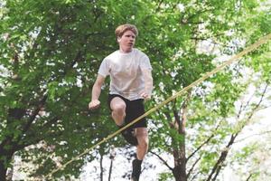 junger Mann balanciert und springt auf Slackline. Mann geht, springt und balanciert auf einem Seil im Park. foto