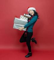 hübsche lächelnde asiatische frau im warmen weihnachtspullover und in der weihnachtsmütze, die geschenkbox als geschenk mit rotem hintergrund für saisonfeierkonzept hält foto