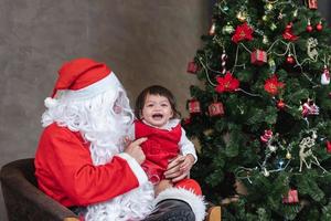 der weihnachtsmann hebt ein glückliches kleines kleinkindbaby hoch und lacht fröhlich mit einem vollständig geschmückten weihnachtsbaum auf dem rücken für das saisonfeierkonzept foto