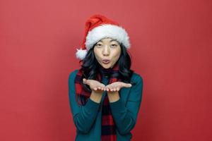 hübsche lächelnde asiatische frau im warmen weihnachtspullover und in der weihnachtsmütze bläst einen grußwunsch auf rotem hintergrund für das saisonfeierkonzept foto
