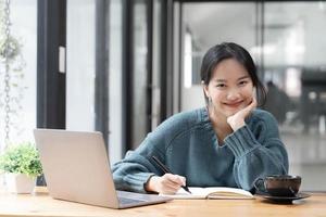 schöne studentin, die online studiert, macht sich notizen auf ihrem laptop, um informationen über ihr lächelndes gesicht und eine glückliche studienhaltung zu sammeln. foto