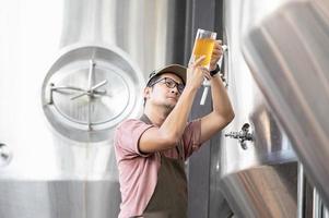 junger asiatischer arbeiter, der die brauereiqualität mit einem glas craft beer inspiziert und das visuelle erscheinungsbild nach der vorbereitung bewertet, während er in einer handwerklichen brauerei arbeitet. foto